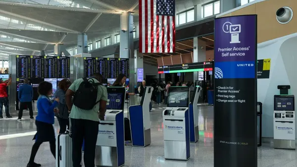 Travelers stand at an airport kiosk with an American flag overhanging.