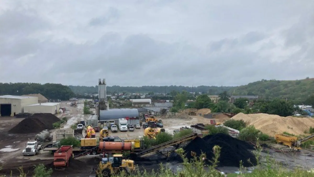 Industrial site shown from elevated angle with piles of sand, dirt and mulch heaped higher than nearby trucks. Various industrial equipment strewn about.