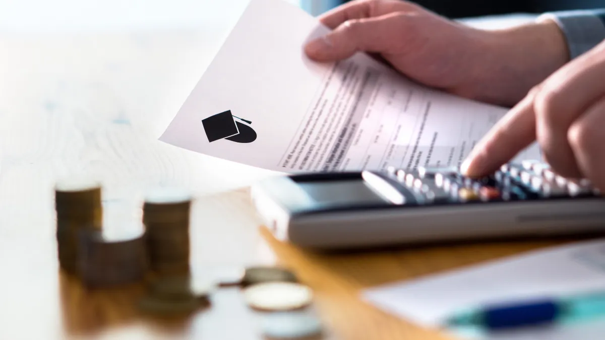 A close up of a pair of hands holding an education invoice and typing on a calculator that sits on a desk alongside a stack of coins and paper and a pen.