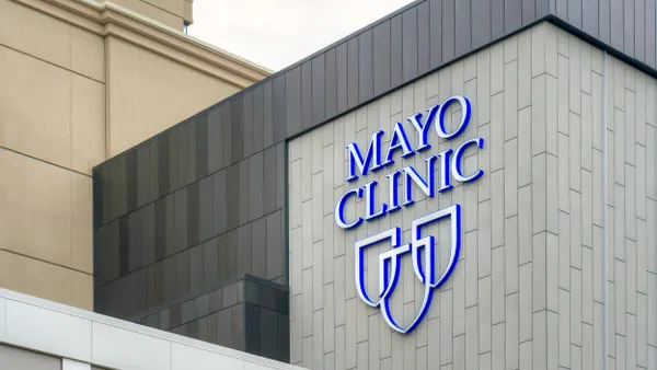Close-up of a modern building with the Mayo Clinic logo in blue and white, displayed on a vertical tiled wall with part of the adjacent structure visible.