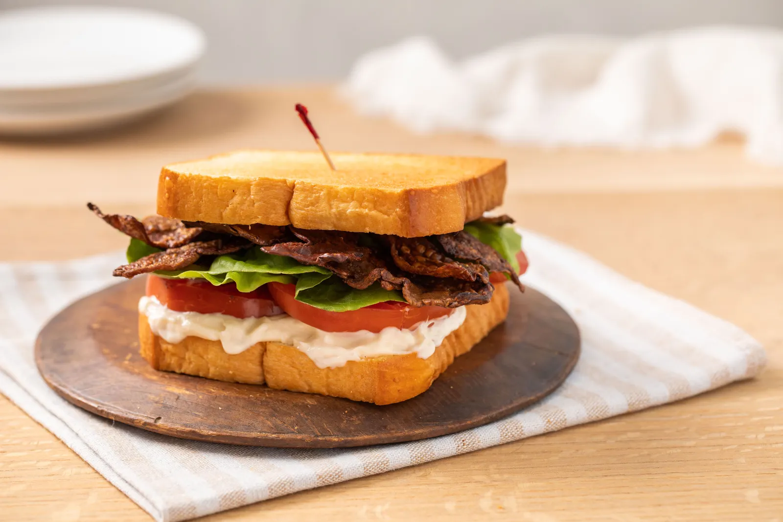A BLT sandwich with lots of mayonnaise made with MyBacon sits on a wooden circle plate on a folded checked cloth napkin on a light wood table.