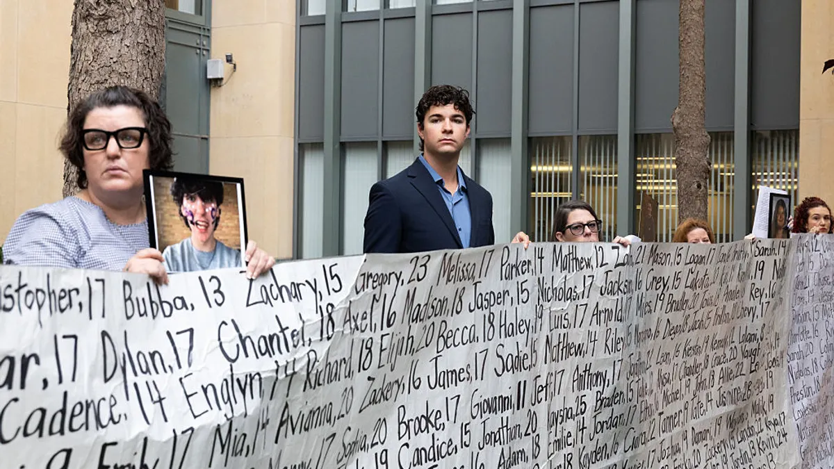 Several people stand outside behind a large banner that lists names and ages of people.