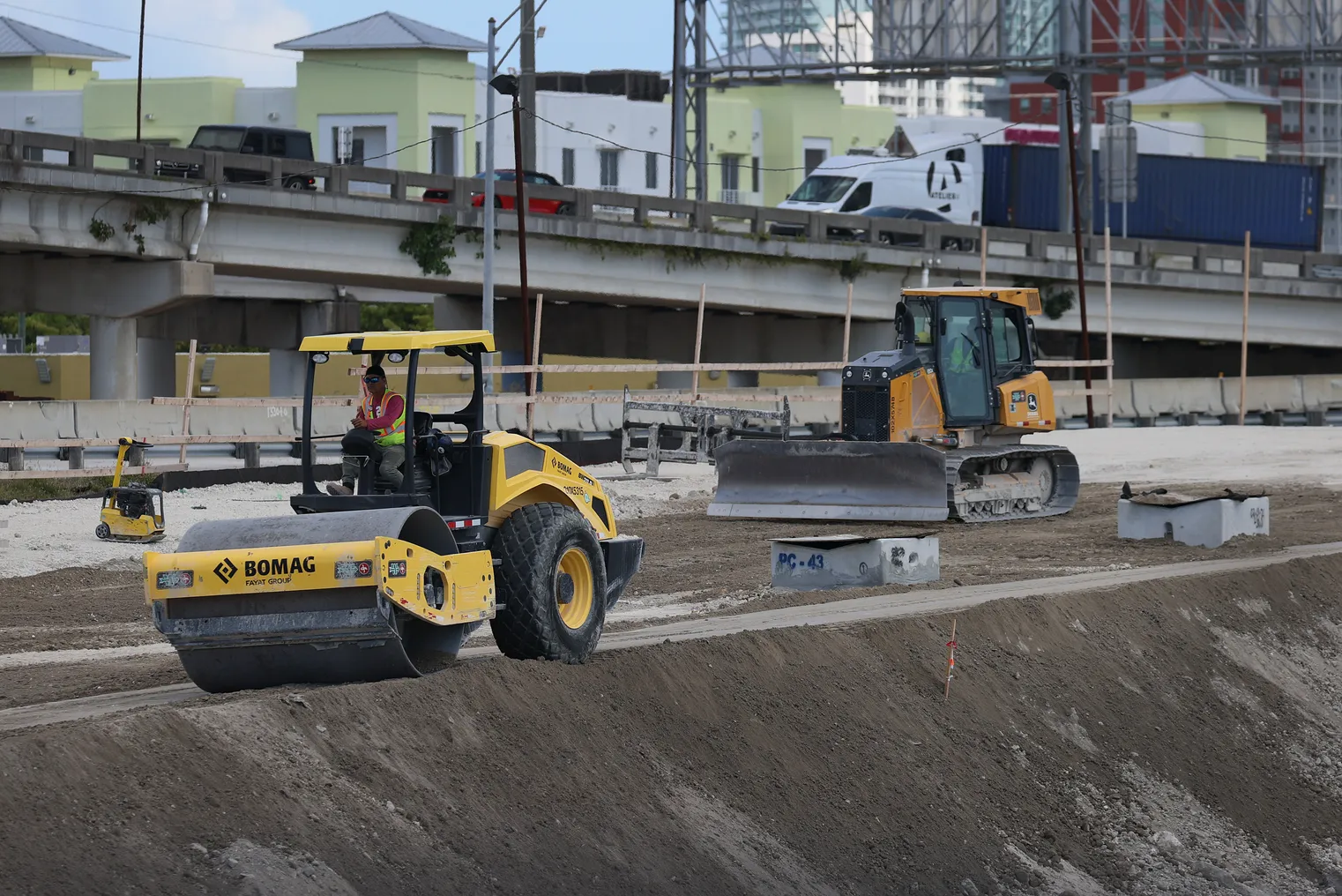 Construction worker in a yellow machine packs dirt on a jobsite with a bridge and more equipment in the background.