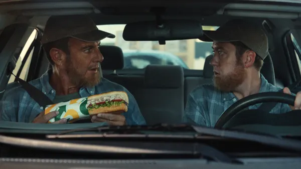 Will Arnett sits alongside a driver in a drive-thru lane while holding up a Subway sandwich