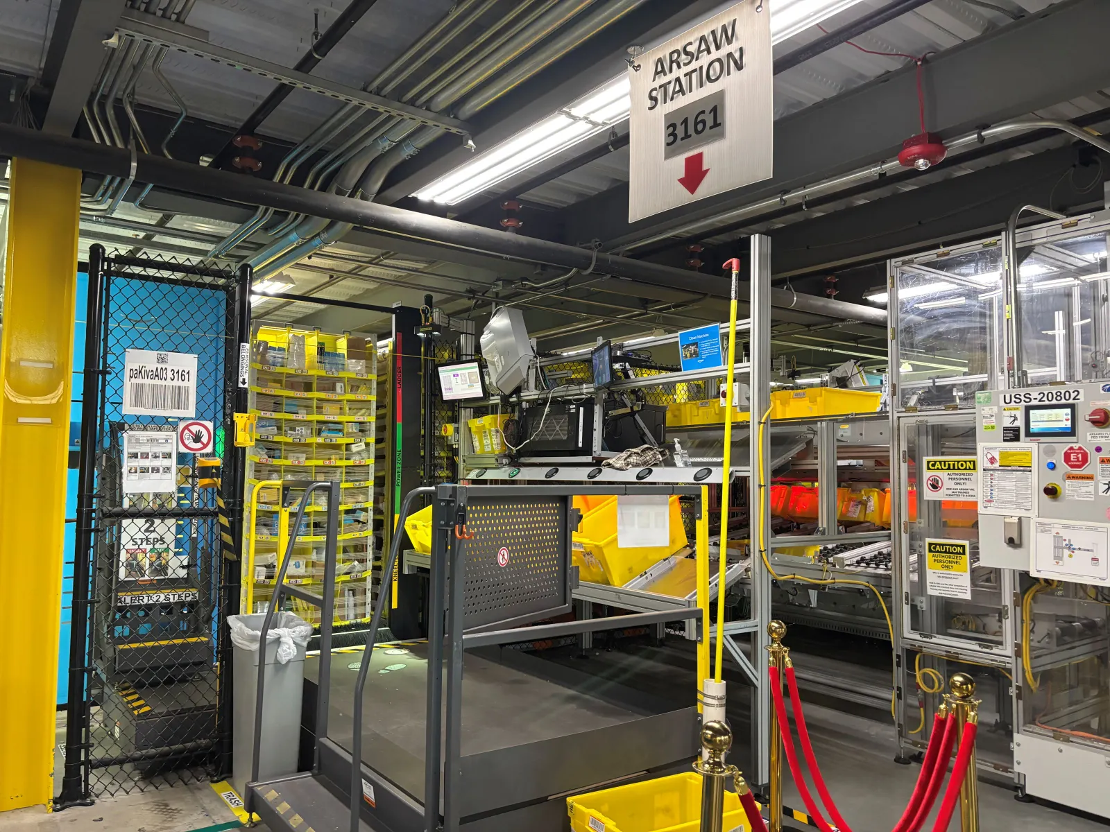 A warehouse workstation with an “ARSAW” sign above, and a shelving unit in the background.
