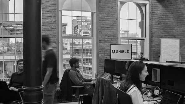 People at work inside a tech firm with the word "shield" displayed on a computer monitor.