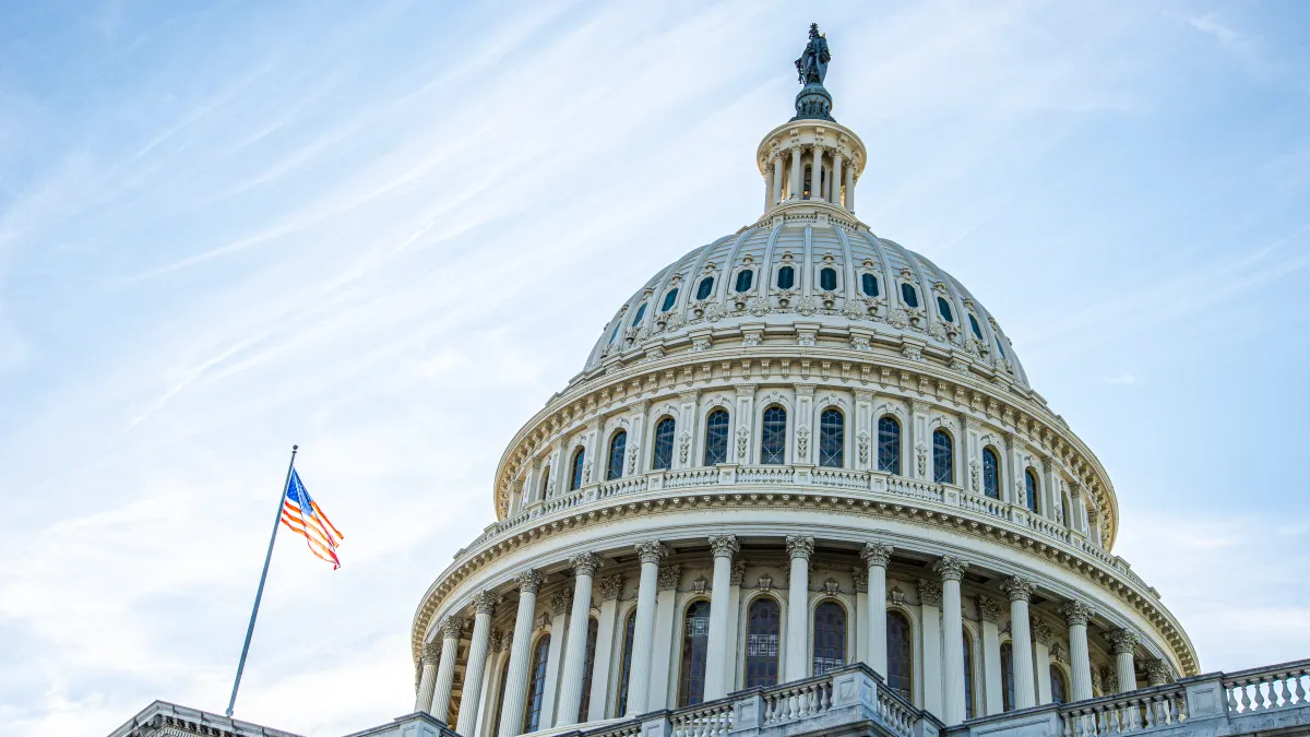 A view of the Capitol Dome from below with an American flag waving in the wind on the left side and blue skies in the background.