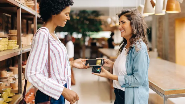 Two women smiling in a store; one is holding a smartphone near a card reader for payment. The scene conveys friendliness and modern shopping.