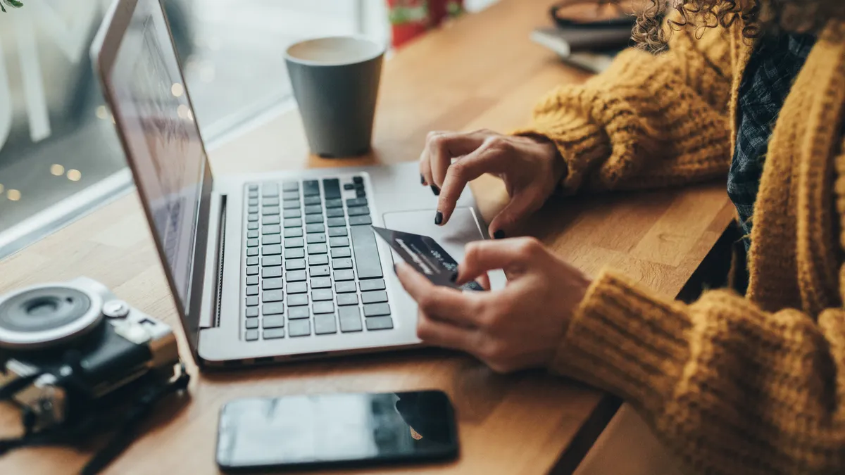 A person with curly hair wearing a sweater holds a credit card as they use a computer.