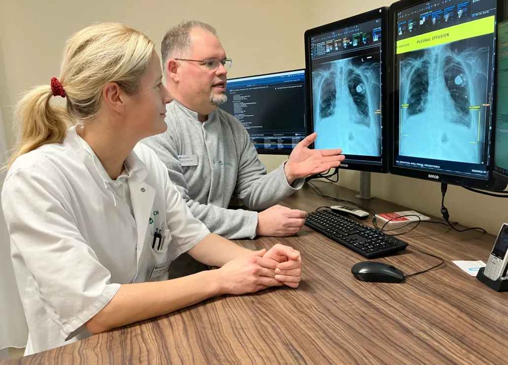 Two people sit in front of three computer monitors showing chest x-rays. One person gestures at the screen.