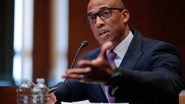 A seated person wearing a suit and glasses gestures while speaking into a microphone.