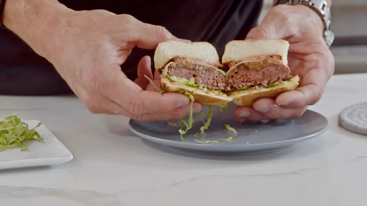 Hands hold a hybrid plant-based and cultivated meat burger cut in half.