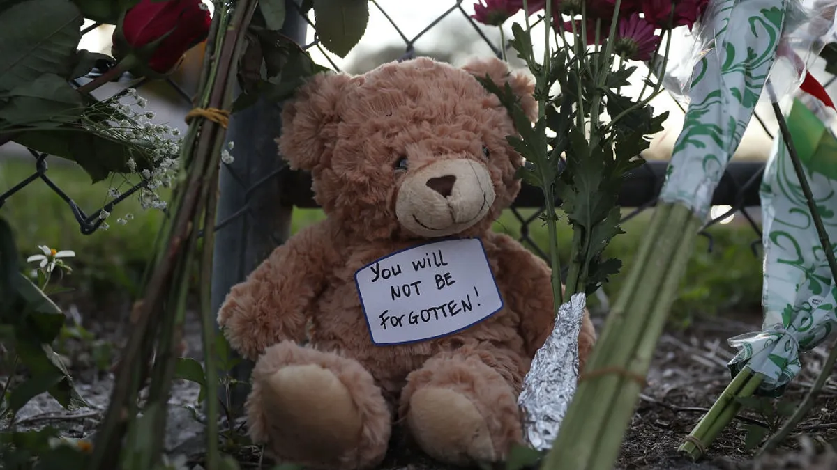 A teddy bear is leaning against a wire fence next to stems of flowers. A note on the teddy bear reads "You will not be forgotten."