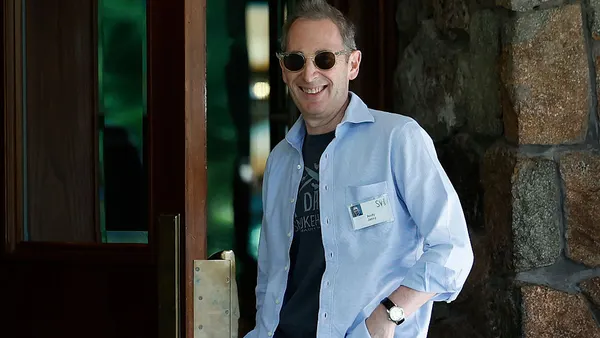 A smiling person in a light blue shirt wears sunglasses, a watch and a nametag.