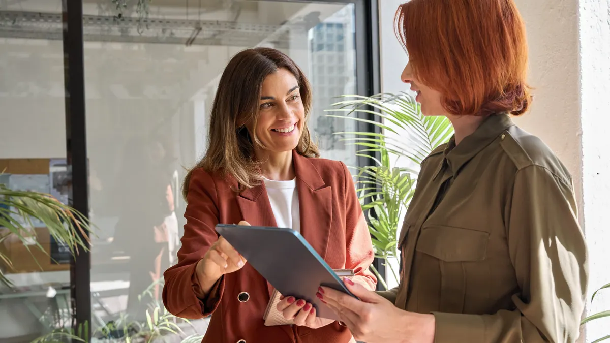 Two happy business women of young and middle age talking standing in creative green office space.