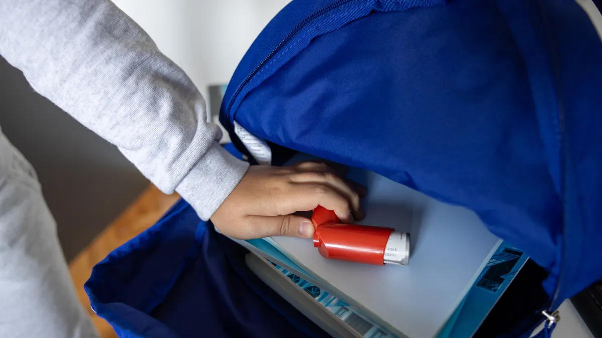 An open backpack on a table shows a laptop and an inhaler. A person's arm is reaching into the backpack.