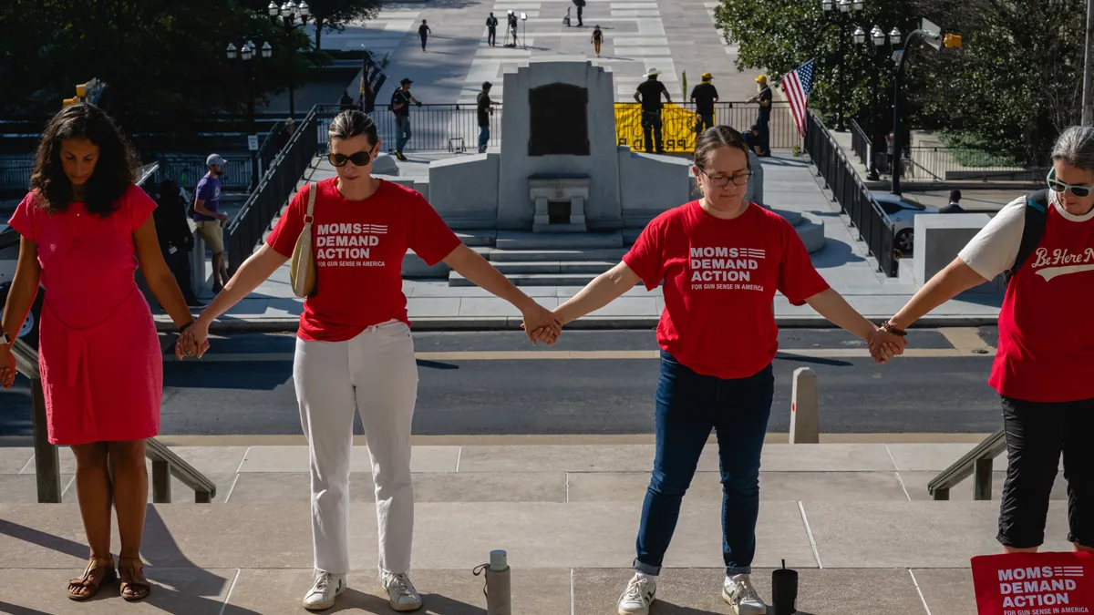 People wearing red t-shirts reading "Moms Demand Action" stand hand-in-hand