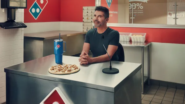 TV personality Simon Cowell, wearing a black T-shirt, sits behind a metal kitchen counter with Domino's branding