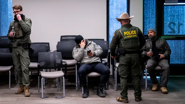 A person wearing a green uniform and the back of a person in a green uniform that says "POLICE" in a waiting room with two people sitting in chairs.