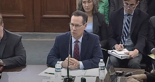 Matthew Perry, CEO of Genesis Healthcare System, wears a navy suit in front of an audience in a Hill briefing room.