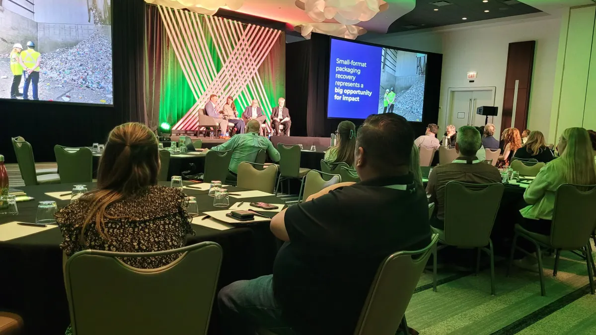 People sit at tables in a large room while observing speakers sitting in chairs on a stage.