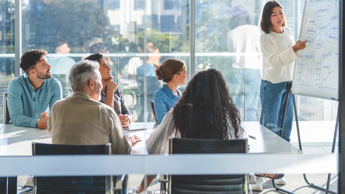 Members of a committee sit at a table and watch a colleague make a presentation on a whiteboard.