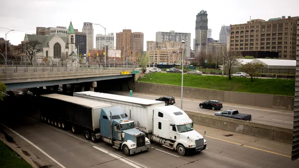 Tractor-trailers emerge from under an overpass while traveling side by side in a city landscape of Detroit.