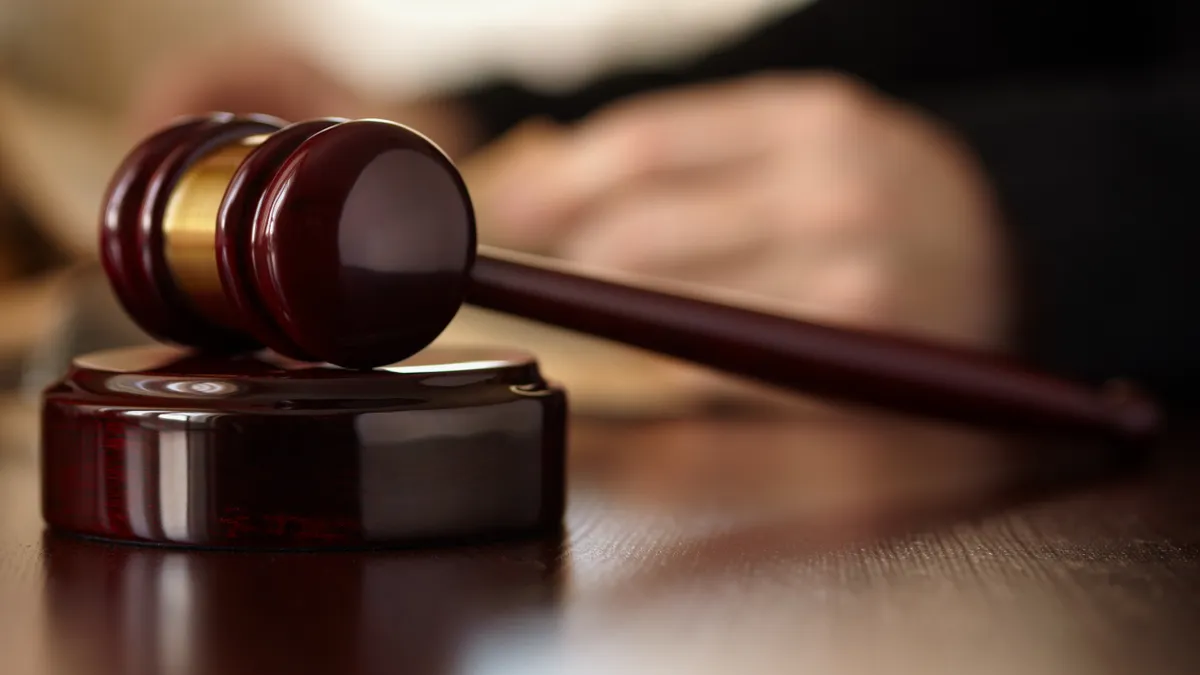 A judge's gavel is shown sitting idle on a thick, cherry stained, perhaps mahogany desk. The judge's hands are visible but out of focus in the background.