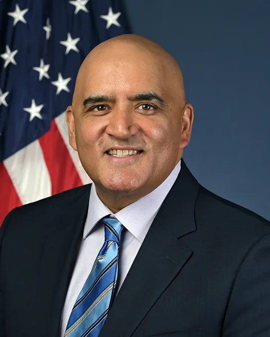 A man in a black suit with a blue tie smiles with an American flag in the background.