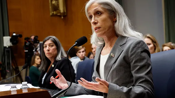 A person in business attire speaks into a microphone while seated at a desk.