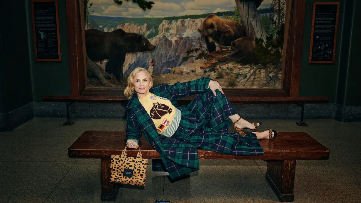 A person reclines on a bench in front of a large painting of three bears. They are wearing a plaid green outfit with a bright yellow sweater and leopard print heels that match their bag.