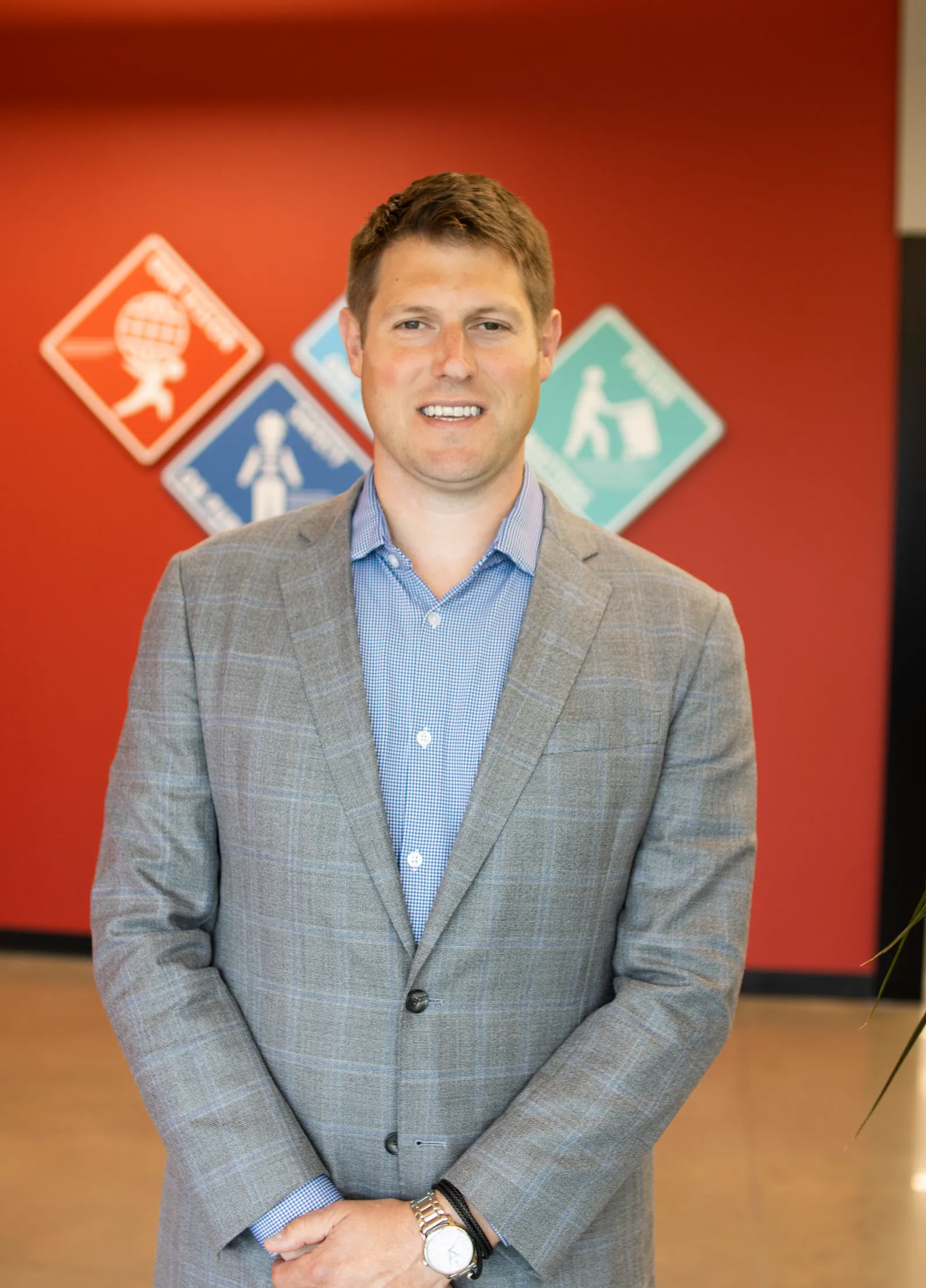 A person in a suit and button-down shirt, with a watch, stands in an office with a red wall.