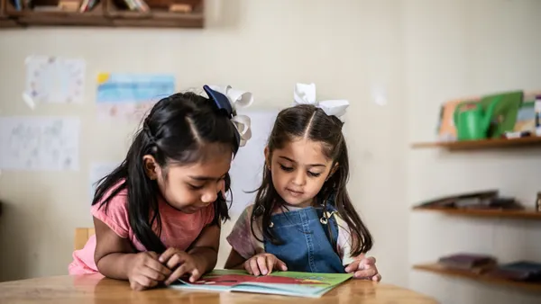 Two young students are in a classroom. They are sitting at a desk and looking at a book together.