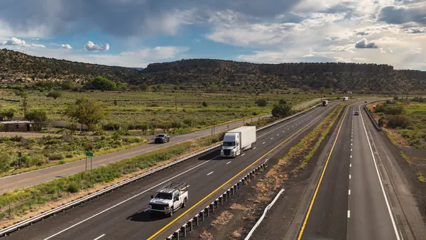 Semi-trucks and pickup trucks on an interstate and the Historic Route 66 with rolling green shrubs, distant sandstone mesas, and cumulus clouds in a bright blue sky in the backdrop.