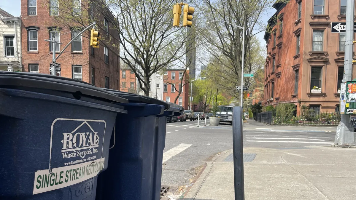 Blue recycling carts in the foreground, with brownstones and Brooklyn streets in the background