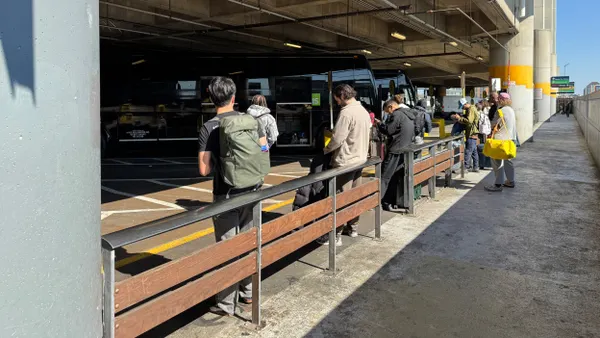 Standing people with luggage lean against a low fence with dark gray buses lined up behind them.