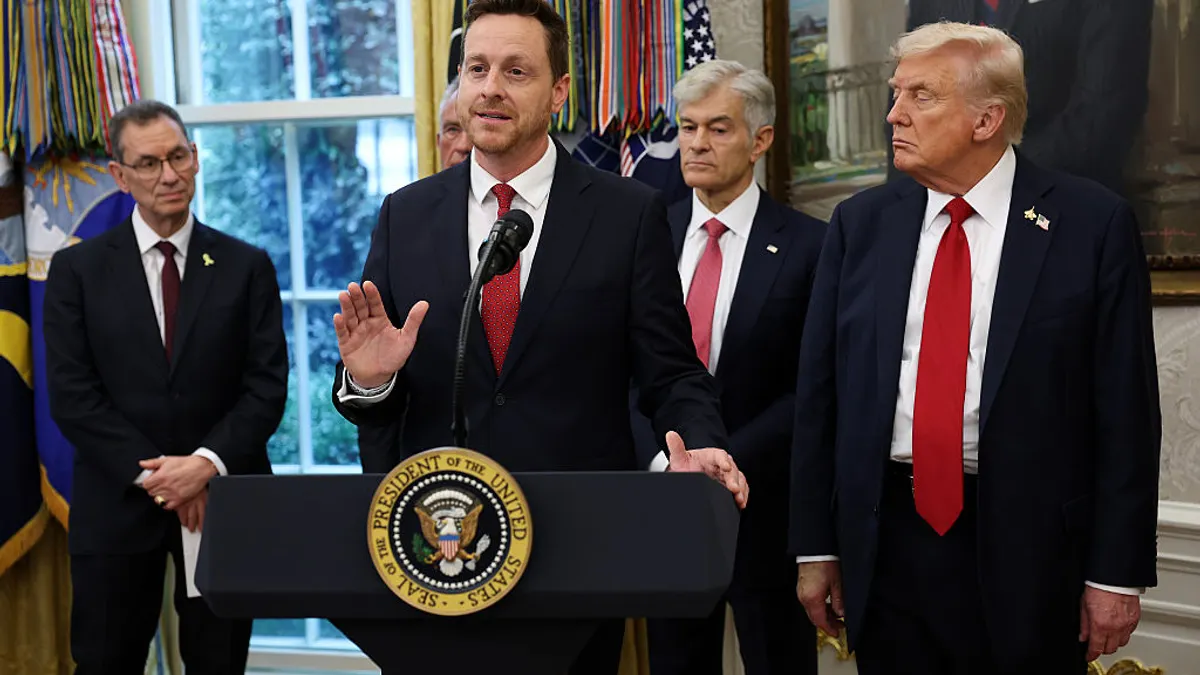 A pan wearing a black suit stands behind a podium flanked by other officials