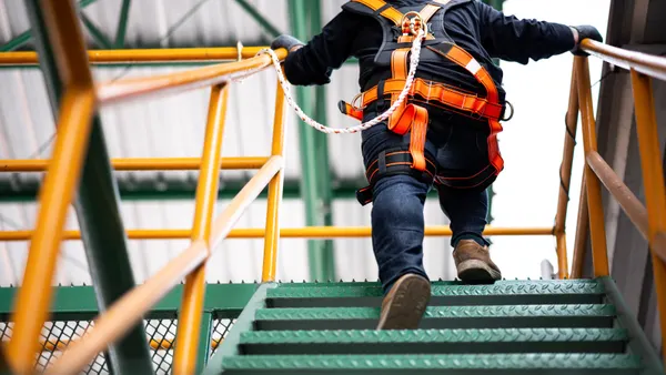 A person wearing safety gear and a safety harness climbs a set of stairs at height, tied off to an anchor point.