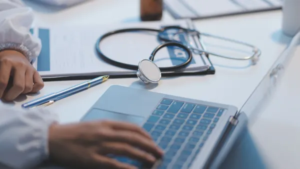 A medical professional rests their hand on a laptop keyboard while a stethoscope sits on the table next to them.