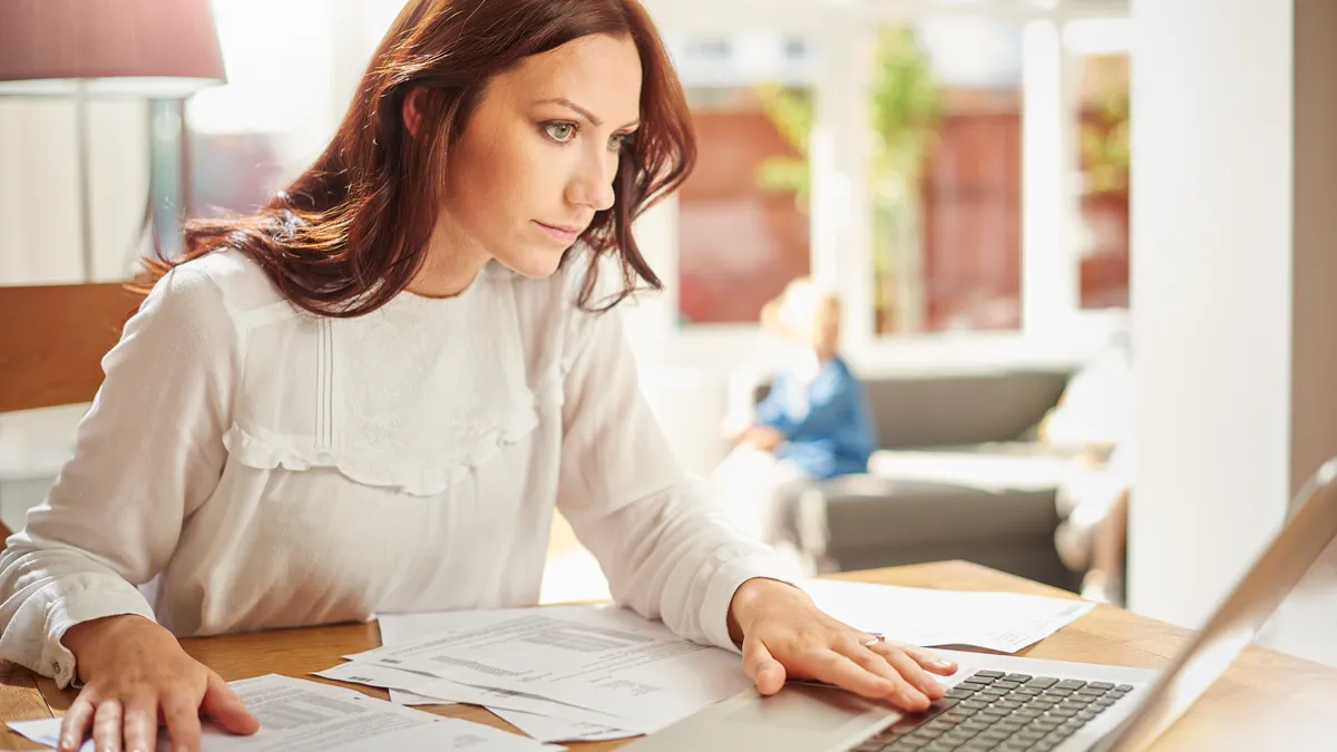 A young woman is sitting at the dining table using her laptop to sort her household bills online.