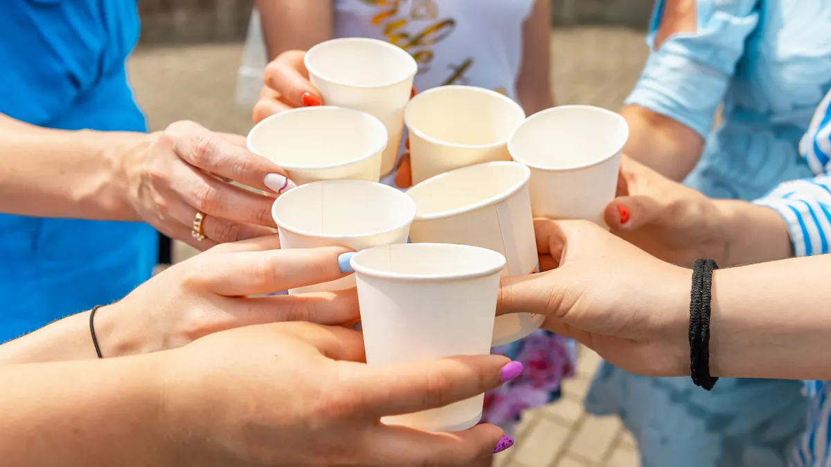 Seven people's hands holding paper cups come together for a toast.