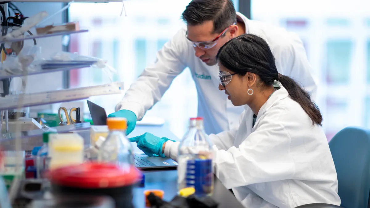 Two researchers in safety goggles and lab coats work in a lab.