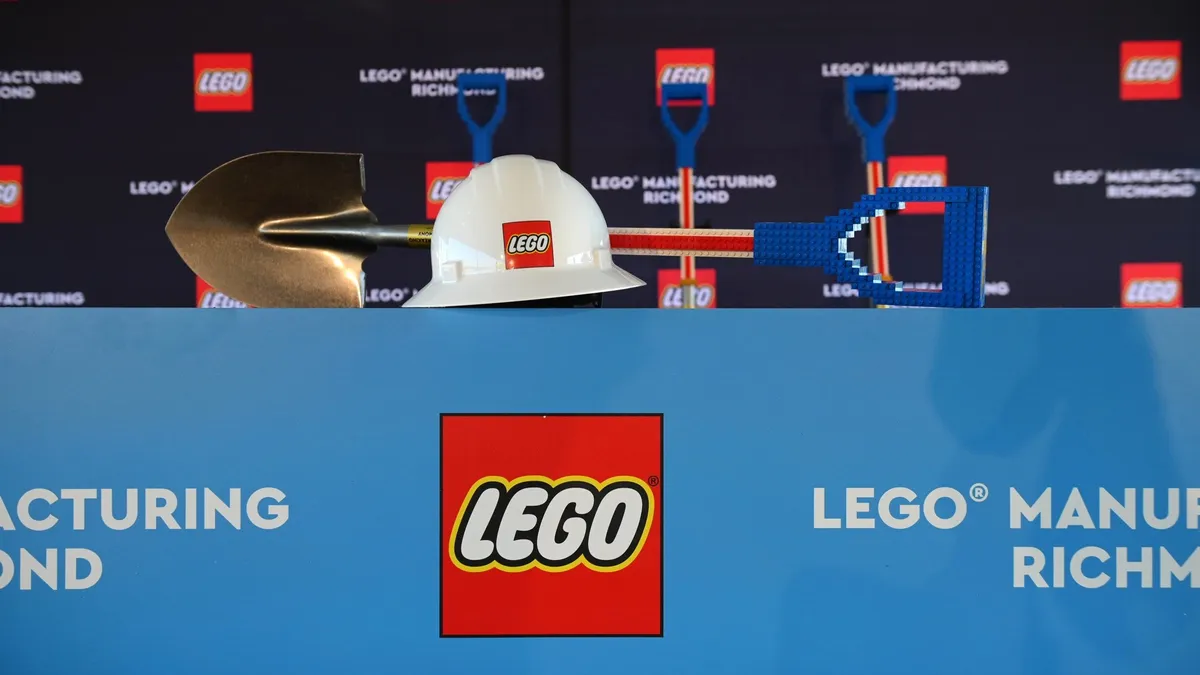 A white hard hat with the red and white Lego logo on the front and a shovel with a blue Lego-brick handle behind it. Both are sitting on top of a light blue Lego sign.