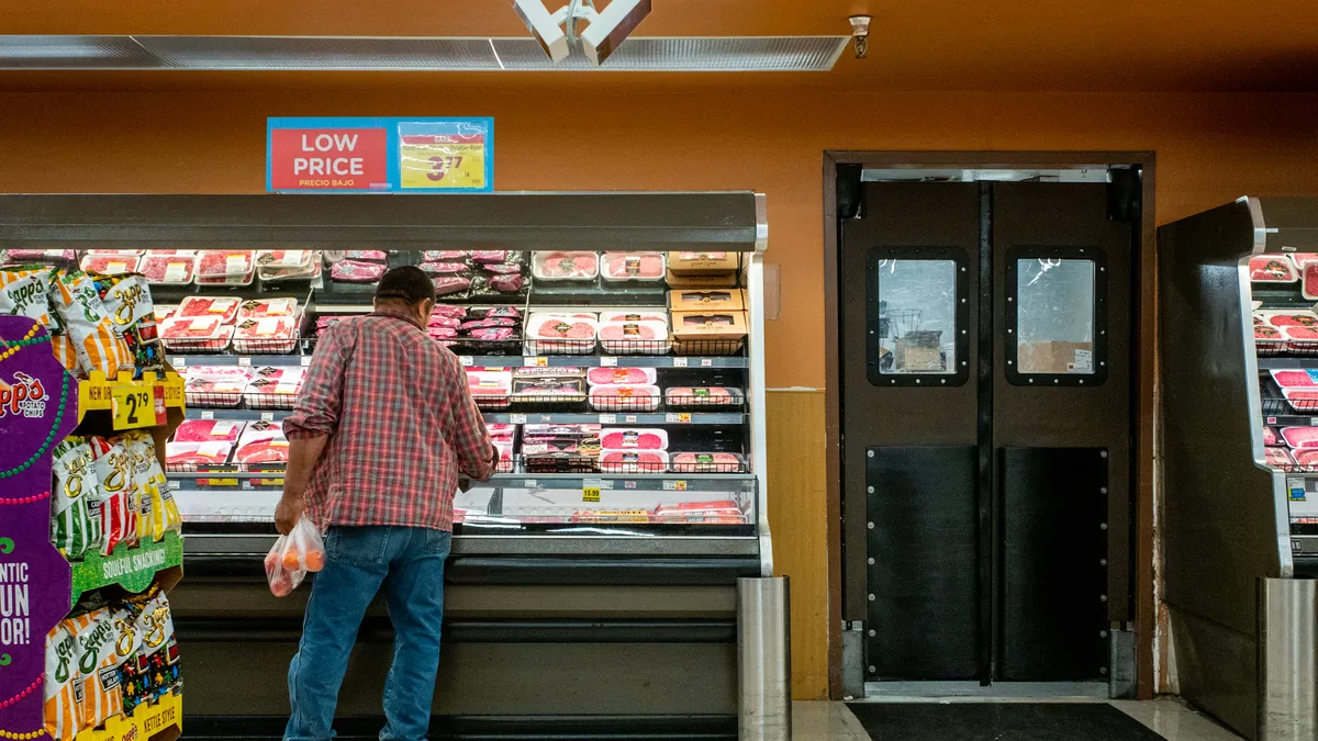 Inside a grocery store, a grocery shopper browses the meat selection where a "low price" sign hangs.