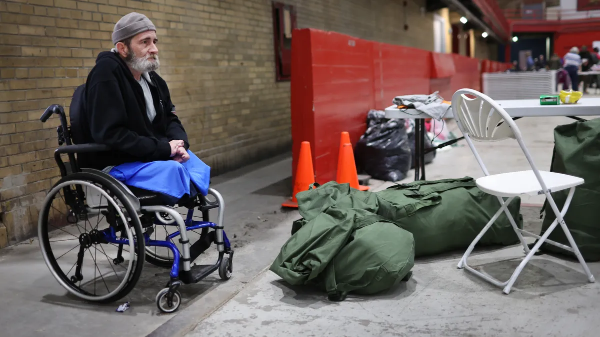 A person in a wheelchair sits next to large bags and a folding chair and table inside.