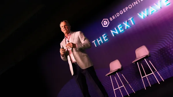 A man in a blazer holding a clicker stands on a stage. The back of the stage is purple and blue and bears the words "Bridgepointe: The Next Wave."