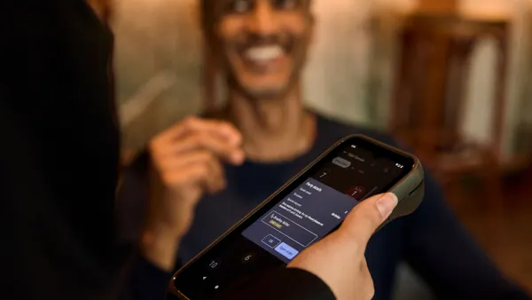 A hand holds a Toast point-of-sale card reader. A customer's face is in the background.