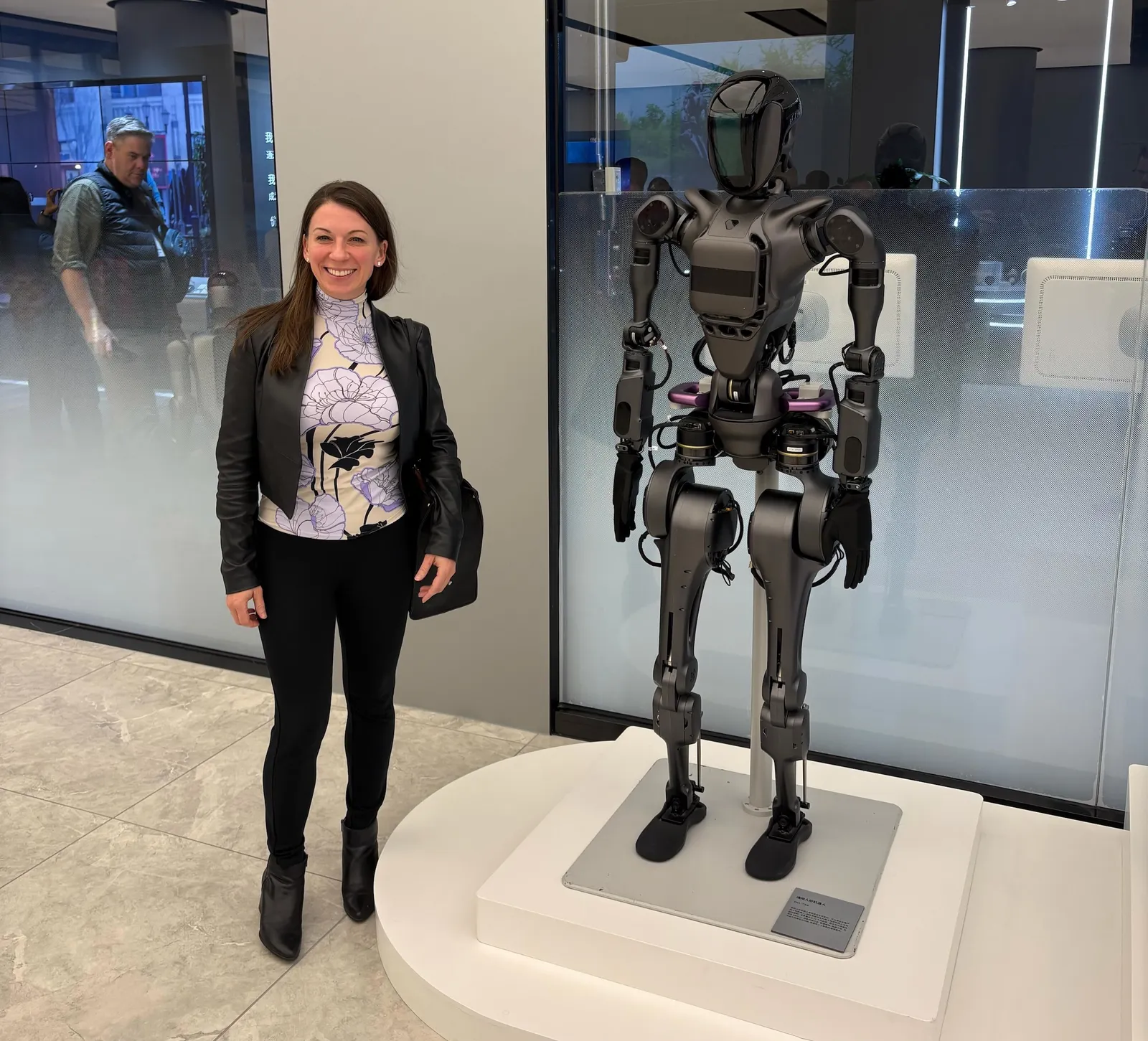 A woman stands next to a robot in a conference center hallway. A window into a conference room is visible behind in the background.