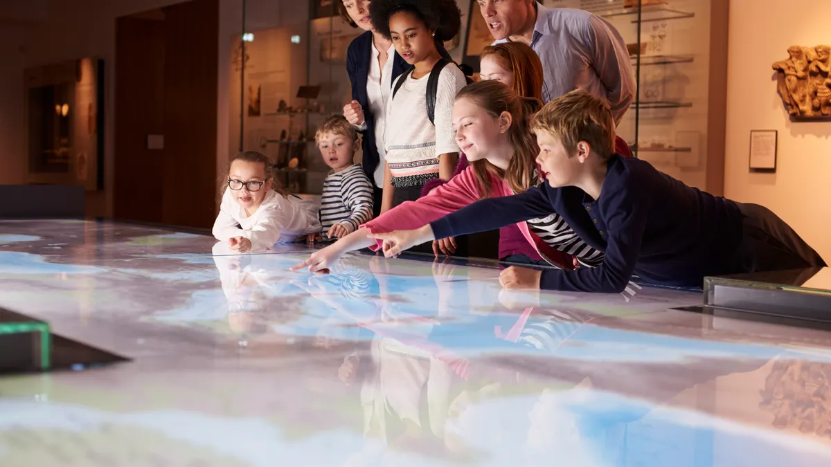A handful of students is looking at and pointing to a light-up table in a room. A few adults are standing beside them