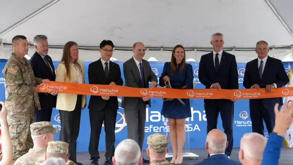 One person in a camouflage uniform, and seven others in business attire, standing on stage holding an orange ribbon.